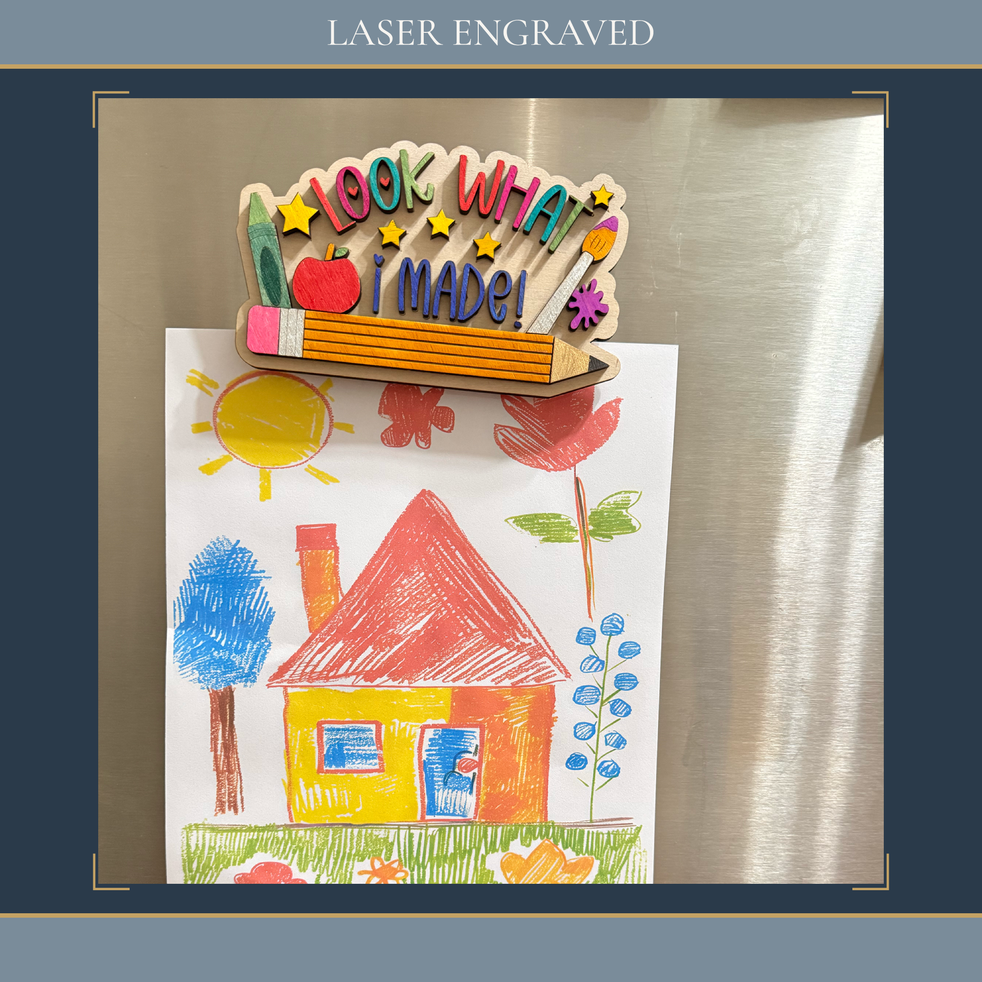 A colorful children's crayon drawing of a house and trees magnetically clipped to a stainless steel refrigerator, with a laser-engraved wooden sign reading 'look what I made!' resting on top. The image is framed with a dark blue border containing the text 'LASER ENGRAVED'.