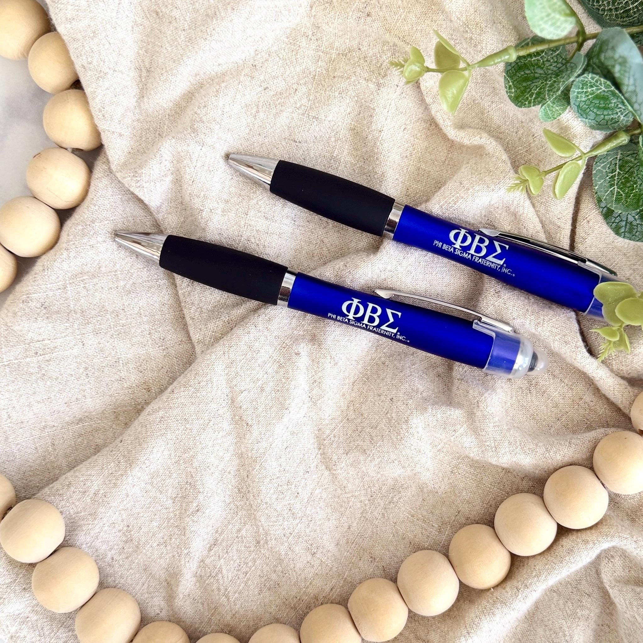 Two blue Phi Beta Sigma engraved pens resting on fabric with greenery and wood beads