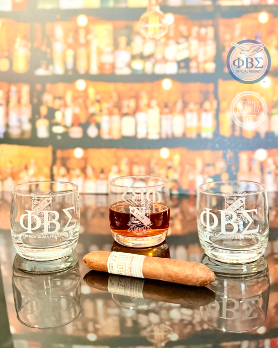Phi Beta Sigma engraved whiskey glasses on a reflective surface with cigar and bar background, official fraternity gift for alumni and members