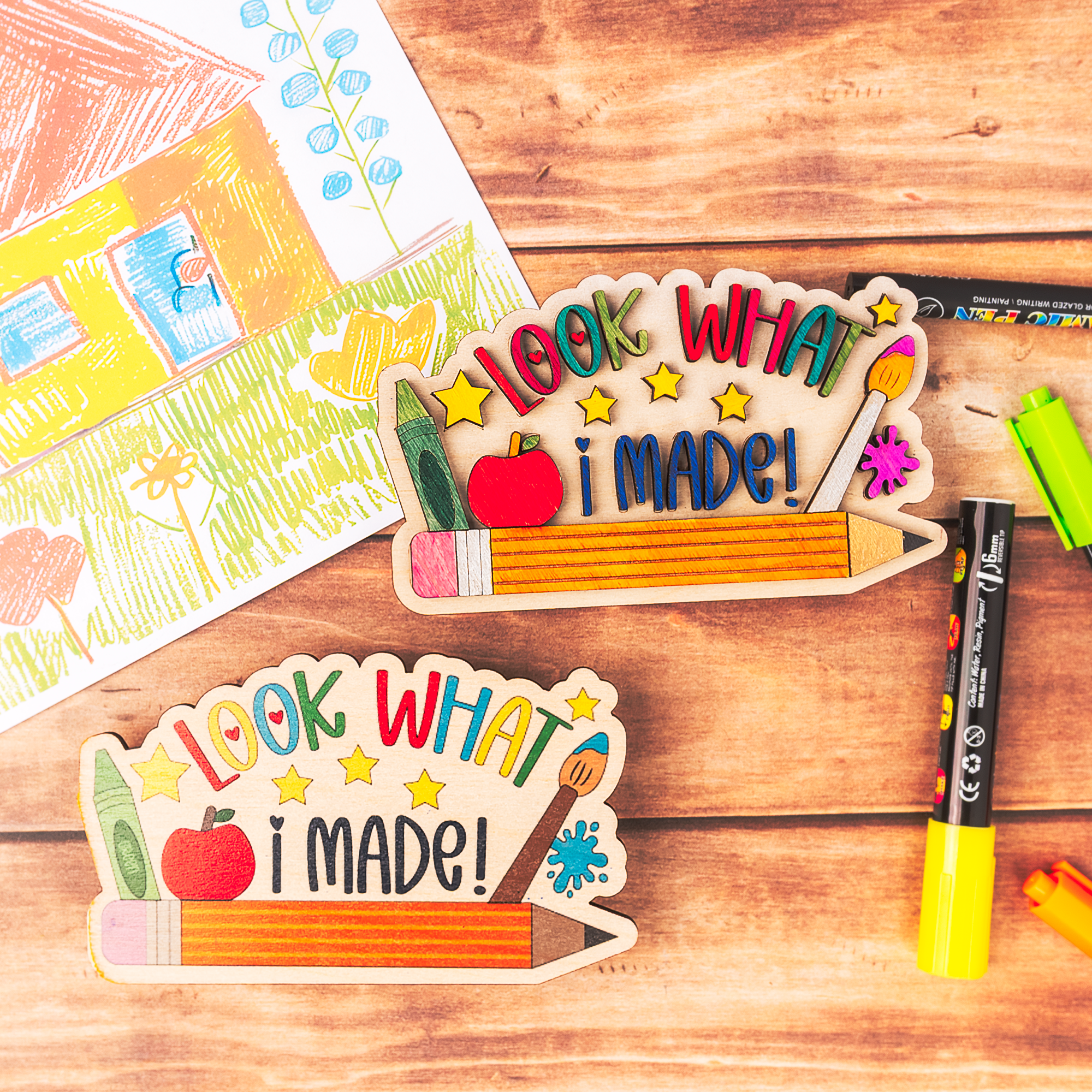 Two wooden school-themed craft plaques reading 'LOOK WHAT I MADE!' are arranged on a wooden tabletop with markers and a child's drawing in the background.