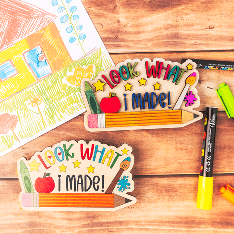 Two wooden school-themed craft plaques reading 'LOOK WHAT I MADE!' are arranged on a wooden tabletop with markers and a child's drawing in the background.