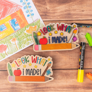 Two wooden craft signs reading 'LOOK WHAT I MADE!' arranged on a wooden tabletop with crayons, markers, and a children's drawing visible around them.