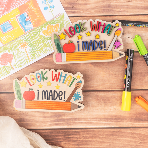 Two wooden craft plaques reading 'LOOK WHAT I MADE!' are arranged on a wooden surface with markers and a children's drawing card nearby. The plaques are shaped like pencils and are decorated with colorful letters, stars, an apple, and art tools.