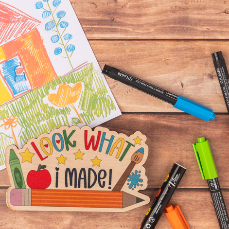 A wooden 'Look What I Made!' UV printed fridge sign displayed on a rustic wooden table alongside various colored markers and a children's drawing.