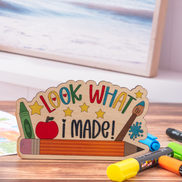 A wooden desk sign shaped like a pencil with the words 'LOOK WHAT I MADE!' in colorful letters, displayed on a wooden surface with crayons and markers nearby.