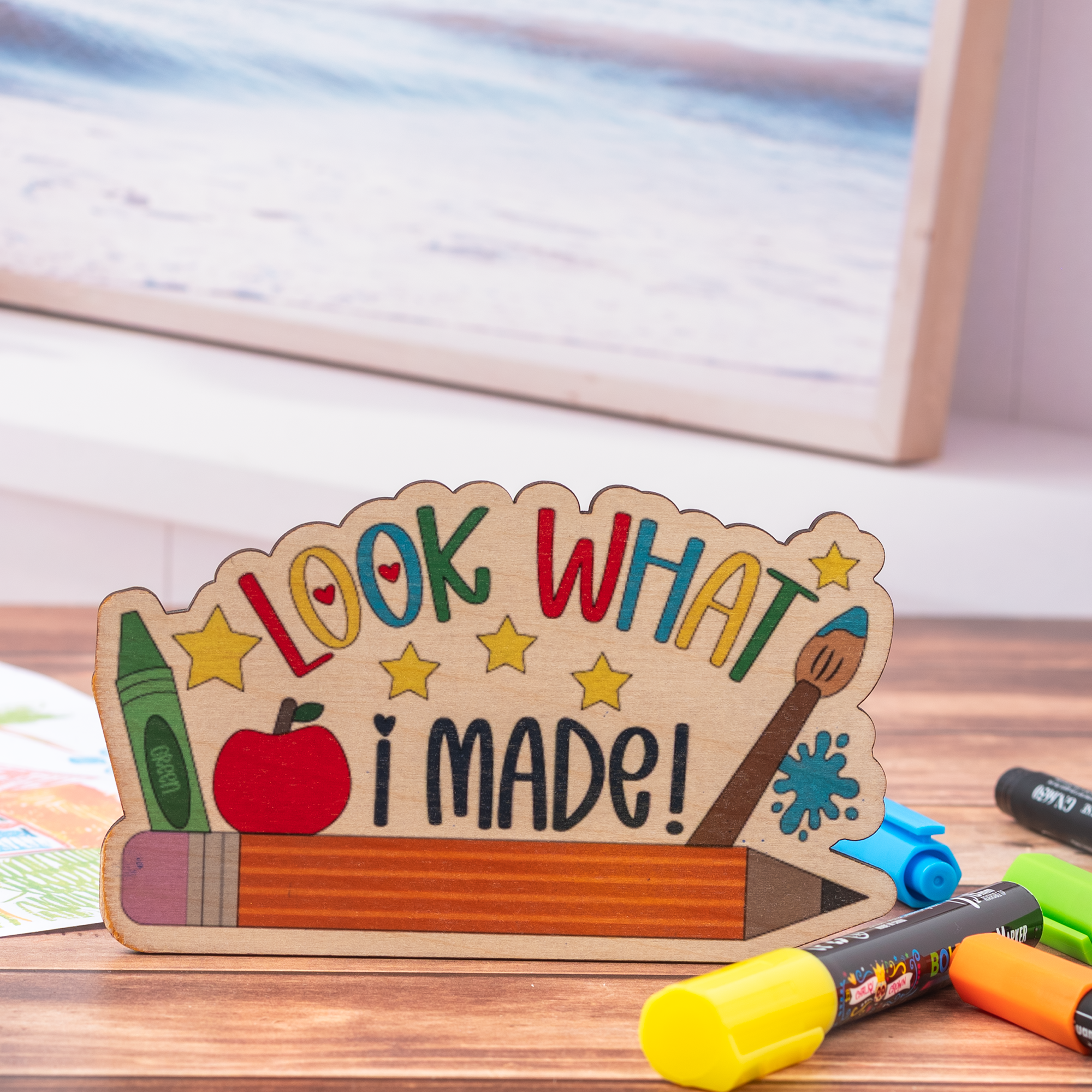 A wooden desk sign shaped like a pencil with the words 'LOOK WHAT I MADE!' in colorful letters, displayed on a wooden surface with crayons and markers nearby.