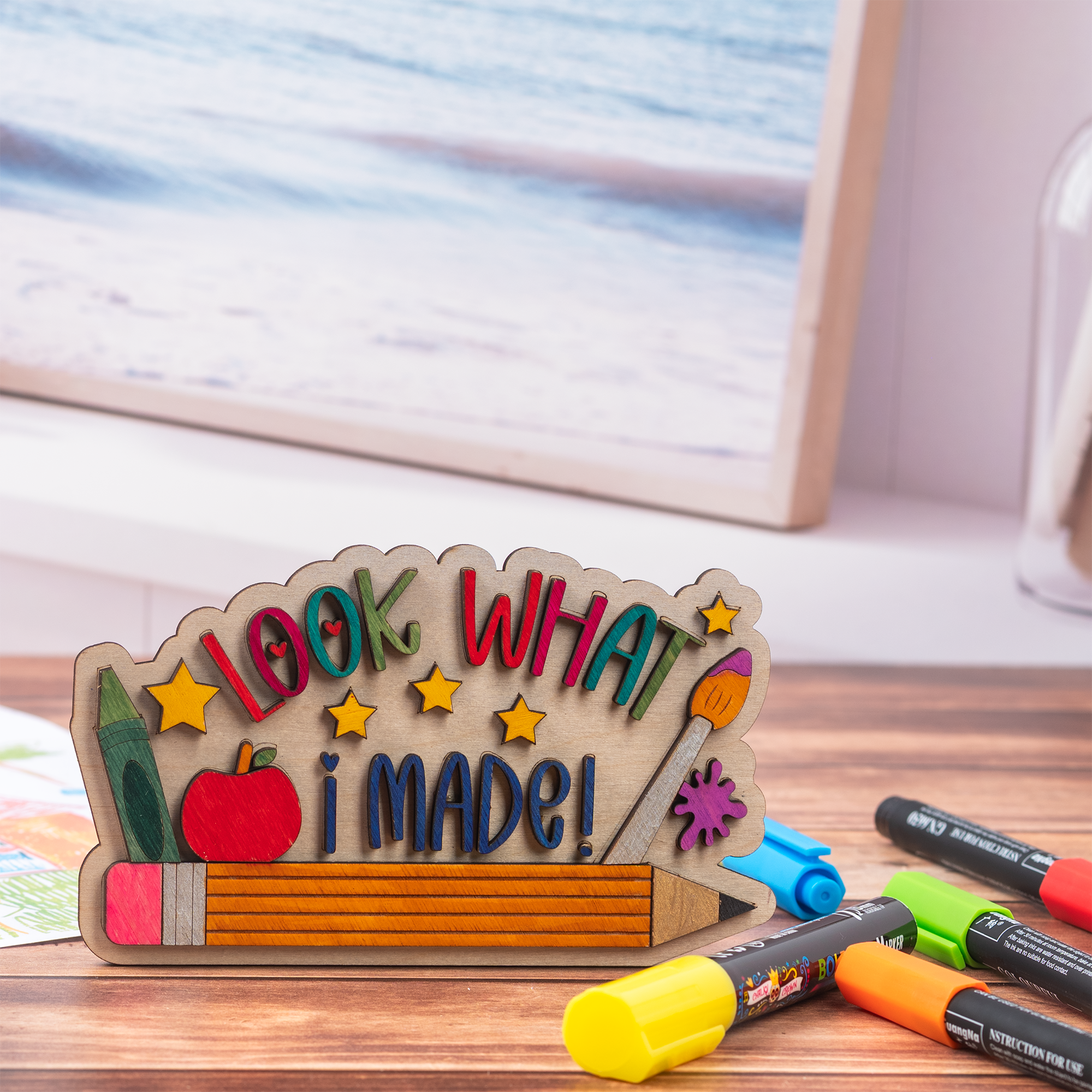 A wooden desk sign with the phrase 'LOOK WHAT I MADE!' in colorful letters, decorated with a pencil, apple, paintbrush, star shapes, and a green eraser. The sign is styled on a wooden tabletop with markers in the foreground and a blurred framed picture in the background.