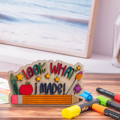 A wooden desk sign with the phrase 'LOOK WHAT I MADE!' in colorful letters, decorated with a pencil, apple, paintbrush, star shapes, and a green eraser. The sign is styled on a wooden tabletop with markers in the foreground and a blurred framed picture in the background.