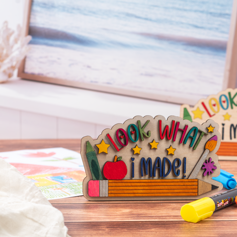 A wooden craft sign that reads 'LOOK WHAT I MADE!' decorated with colorful raised letters, stars, a pencil, an apple, and paintbrushes, displayed on a wooden table with craft supplies in a blurred room background.