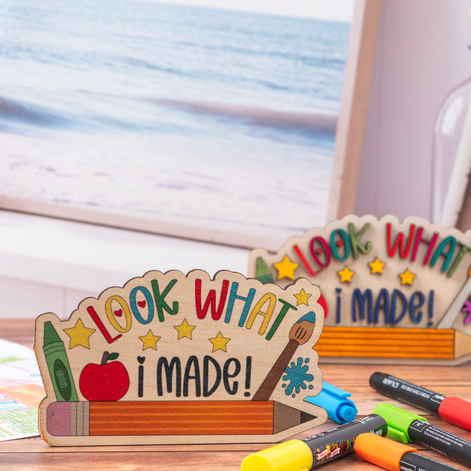A wooden craft display sign reading 'LOOK WHAT i MADE!' with school-themed illustrations, placed on a tabletop near a window with an ocean view. Other markers and another similar sign are visible in the background.